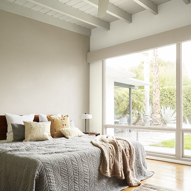 A bedroom painted in neutral hues and white-painted shiplap ceiling with exposed beams, ceiling fan, and a floor-to-ceiling window wall with views of desert landscaping. A bedroom painted in neutral hues and white-painted shiplap ceiling with exposed beams, ceiling fan, and a floor-to-ceiling window wall with views of desert landscaping.