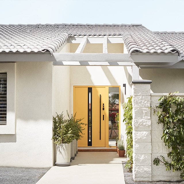 A sunny home exterior painted in off-white with a yellow front door, cacti and other dessert landscaping. A sunny home exterior painted in off-white with a yellow front door, cacti and other dessert landscaping.