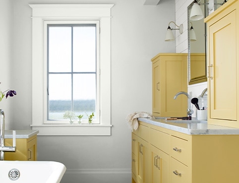 A modern farmhouse style bathroom with off-white walls, white window trim, and soft yellow painted cabinets with silver handles. A modern farmhouse style bathroom with off-white walls, white window trim, and soft yellow painted cabinets with silver handles.