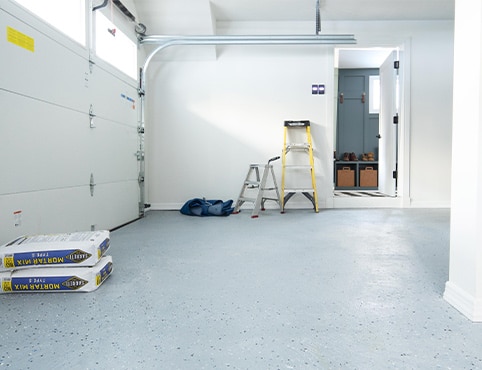 A garage with gray floor and white walls, an assortment of ladders, cement, and a cabinet. A garage with gray floor and white walls, an assortment of ladders, cement, and a cabinet.