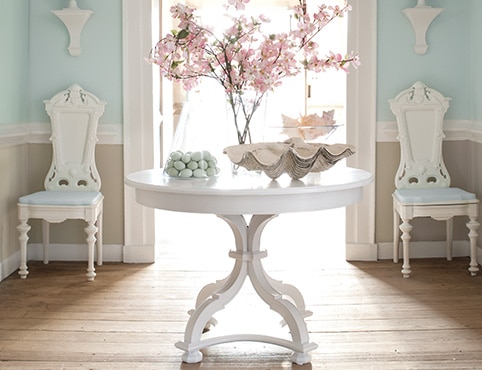 An entryway with a white-painted table with floral decor, two white chairs, and light blue walls, white trim, and beige wainscoting. An entryway with a white-painted table with floral decor, two white chairs, and light blue walls, white trim, and beige wainscoting.