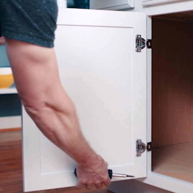 A person holding a screwdriver and removing a white painted kitchen cabinet door from its hinges. A person holding a screwdriver and removing a white painted kitchen cabinet door from its hinges.