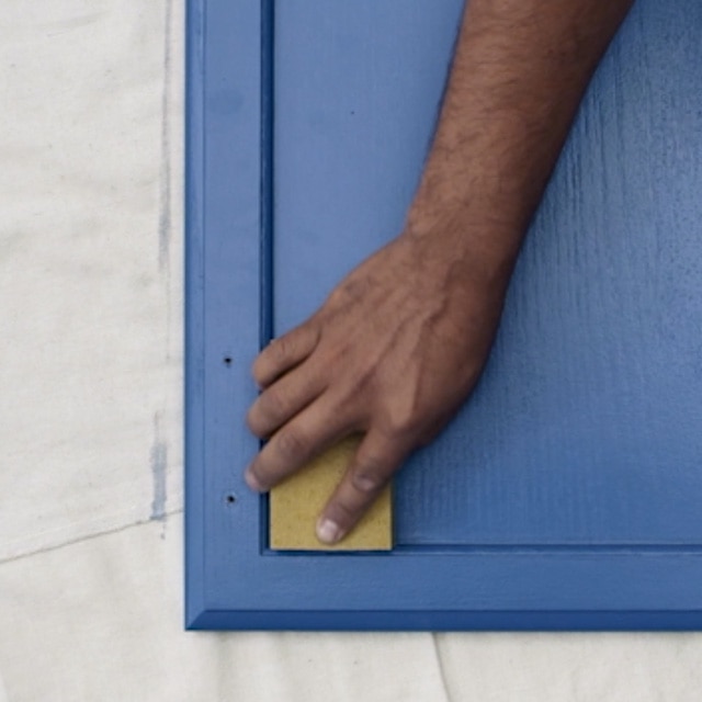 A person sanding a deep blue painted kitchen cabinet door on a white drop cloth between the second recoat. A person sanding a deep blue painted kitchen cabinet door on a white drop cloth between the second recoat.
