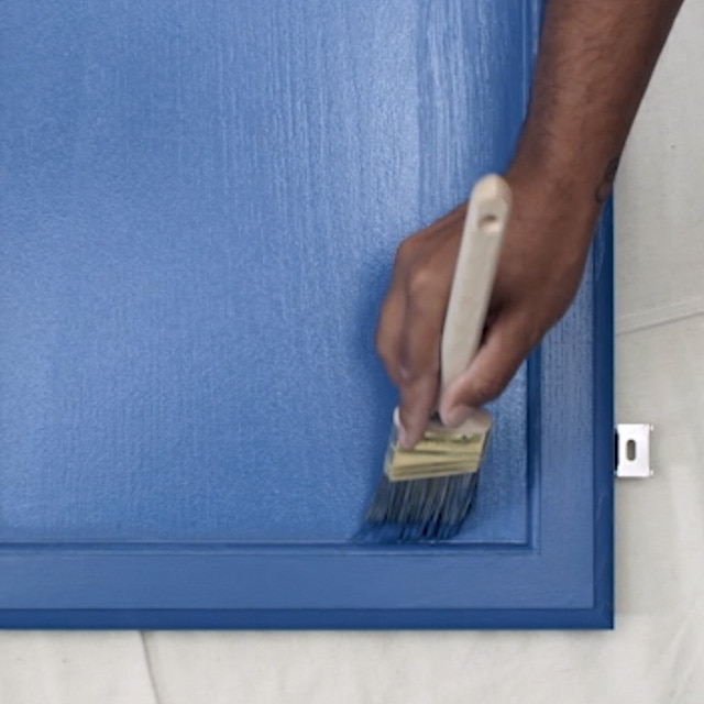 A person brushing deep blue paint onto a kitchen cabinet door that is lying flat on a white drop cloth. A person brushing deep blue paint onto a kitchen cabinet door that is lying flat on a white drop cloth.