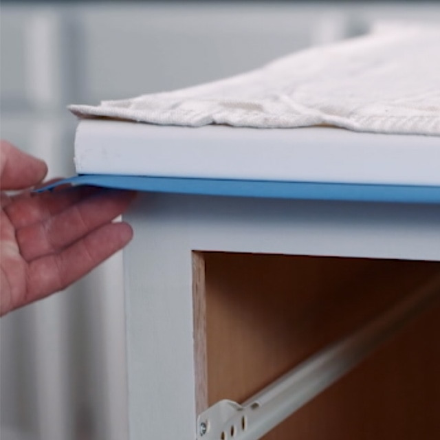 A person applying painter’s tape between the top of a kitchen cabinet frame and the countertop. A person applying painter’s tape between the top of a kitchen cabinet frame and the countertop.