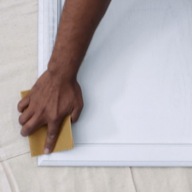 A person sanding a white painted kitchen cabinet door that is lying flat on a white drop cloth. A person sanding a white painted kitchen cabinet door that is lying flat on a white drop cloth.