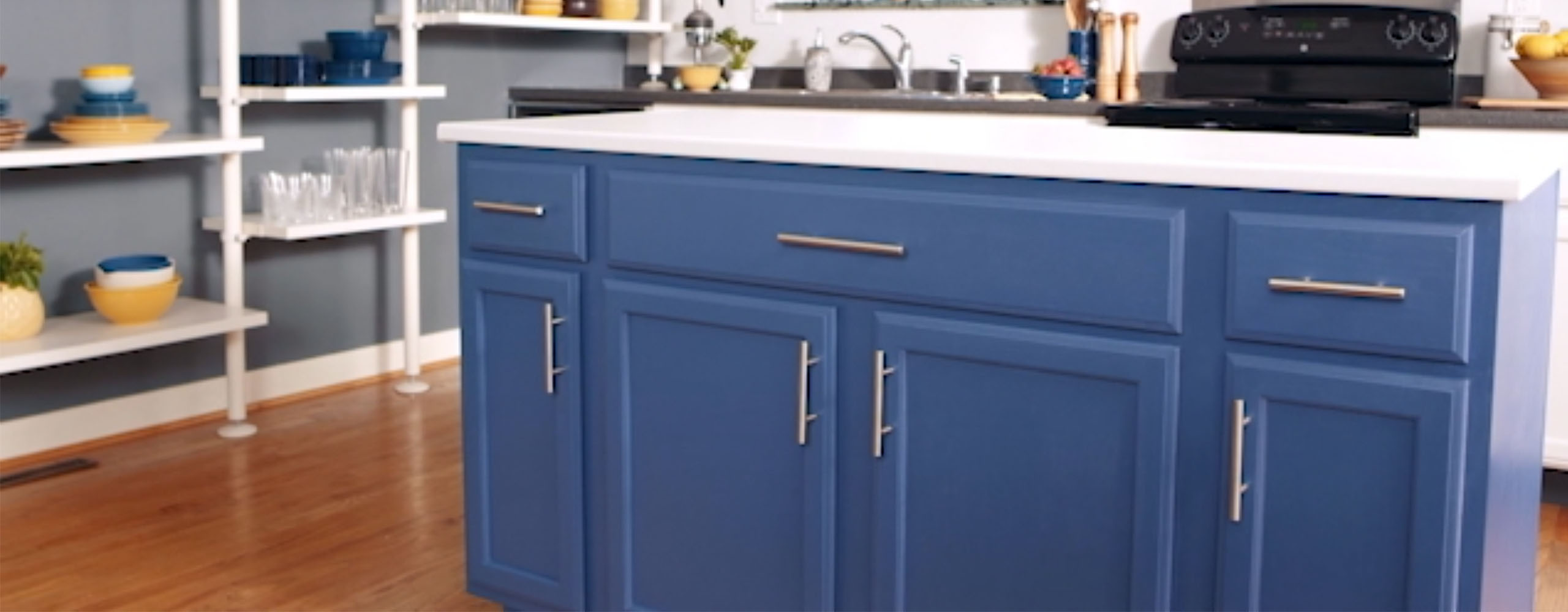 This bright kitchen features a gray-painted side wall with white open shelves, an island with deep blue-painted cabinets and a white countertop, and a white back wall with a window. This bright kitchen features a gray-painted side wall with white open shelves, an island with deep blue-painted cabinets and a white countertop, and a white back wall with a window.