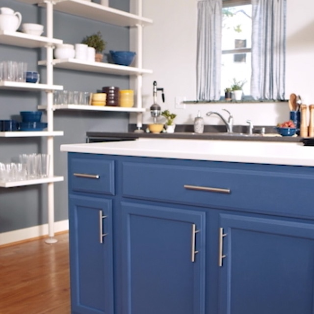 This bright kitchen features a gray-painted side wall with white open shelves, an island with deep blue-painted cabinets and a white countertop, and a white back wall with a window. This bright kitchen features a gray-painted side wall with white open shelves, an island with deep blue-painted cabinets and a white countertop, and a white back wall with a window.