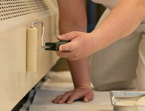 A person rolling off-white paint onto a baseboard heater. A person rolling off-white paint onto a baseboard heater.