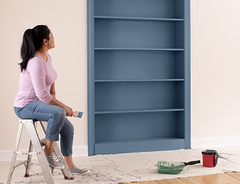 A homeowner sits on a step ladder in a pink-painted room, observing her freshly painted bookcase in a grayish blue. A homeowner sits on a step ladder in a pink-painted room, observing her freshly painted bookcase in a grayish blue.