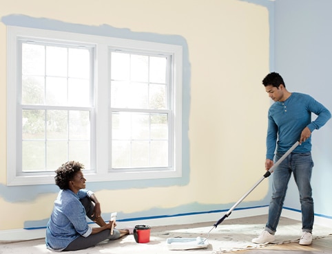 Two homeowners use a paint roller and a paintbrush to paint their beige living room blue, featuring white windows. Two homeowners use a paint roller and a paintbrush to paint their beige living room blue, featuring white windows.