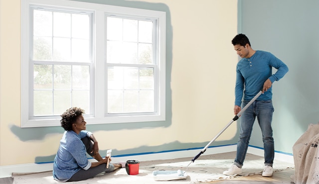 Two homeowners use a paint roller and a paintbrush to paint their beige living room blue, featuring white windows.