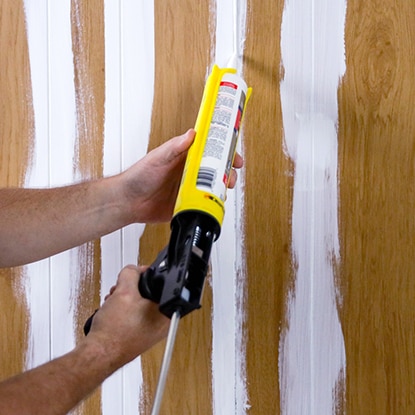 A homeowner uses caulk to fill grooves in wood paneling for a smooth finish. A homeowner uses caulk to fill grooves in wood paneling for a smooth finish.