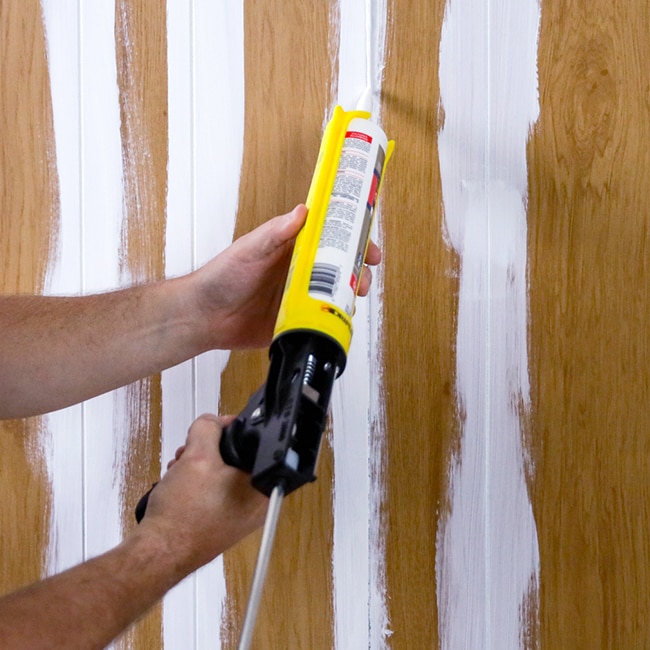 A homeowner uses caulk to fill grooves in wood paneling for a smooth finish. A homeowner uses caulk to fill grooves in wood paneling for a smooth finish.
