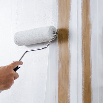 A homeowner rolls paint onto a wood paneled wall. A homeowner rolls paint onto a wood paneled wall.