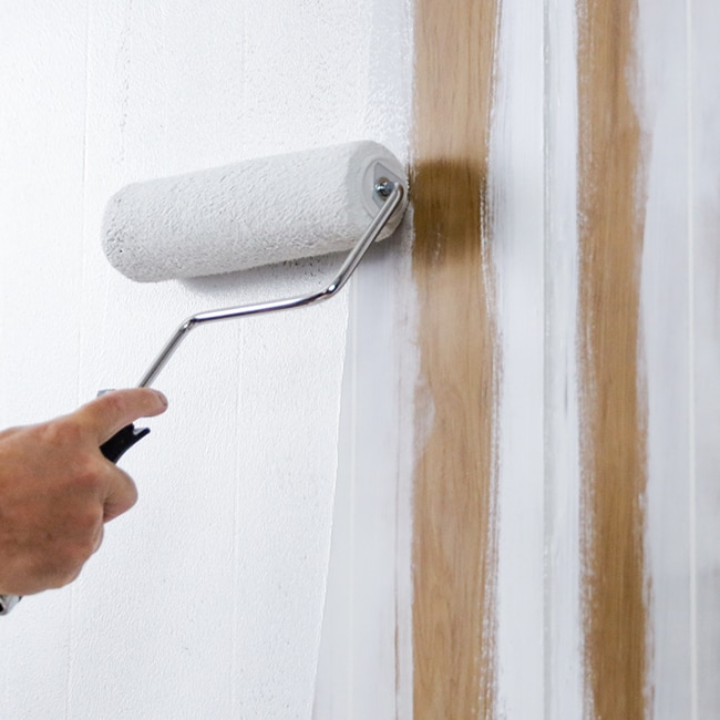 A homeowner rolls paint onto a wood paneled wall. A homeowner rolls paint onto a wood paneled wall.