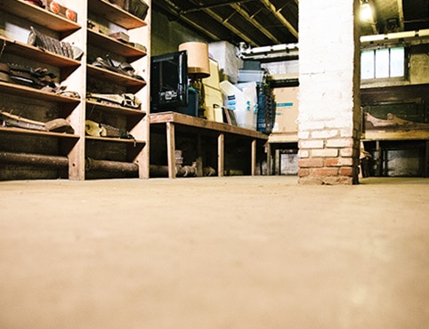 A concrete floor in a garage that needs to be sealed. A brick column is in the middle of the room, and wooden shelves line the walls. A concrete floor in a garage that needs to be sealed. A brick column is in the middle of the room, and wooden shelves line the walls.
