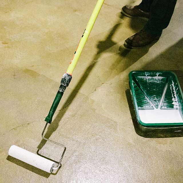 A homeowner applies sealer to a floor using a paint roller. A green painting tray is on the floor. A homeowner applies sealer to a floor using a paint roller. A green painting tray is on the floor.
