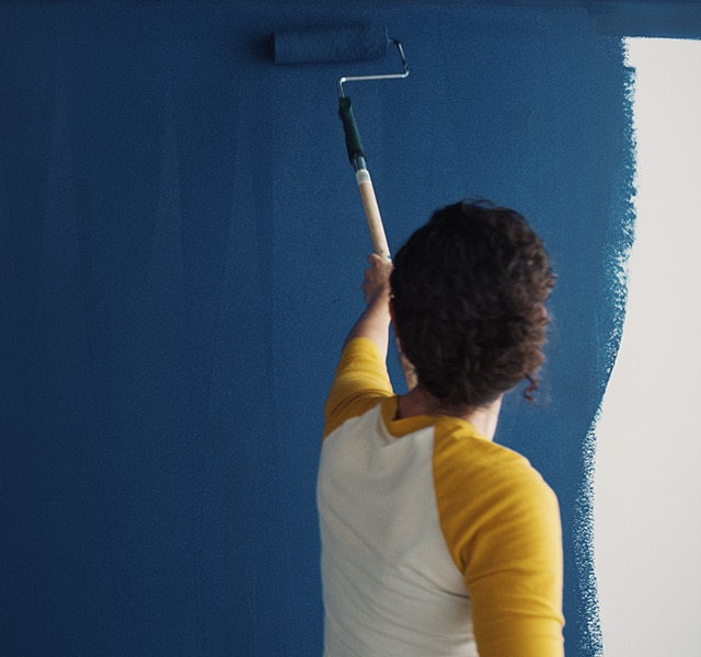 A homeowner uses a roller to paint Symphony Blue 2060-10 on a wall. A homeowner uses a roller to paint Symphony Blue 2060-10 on a wall.