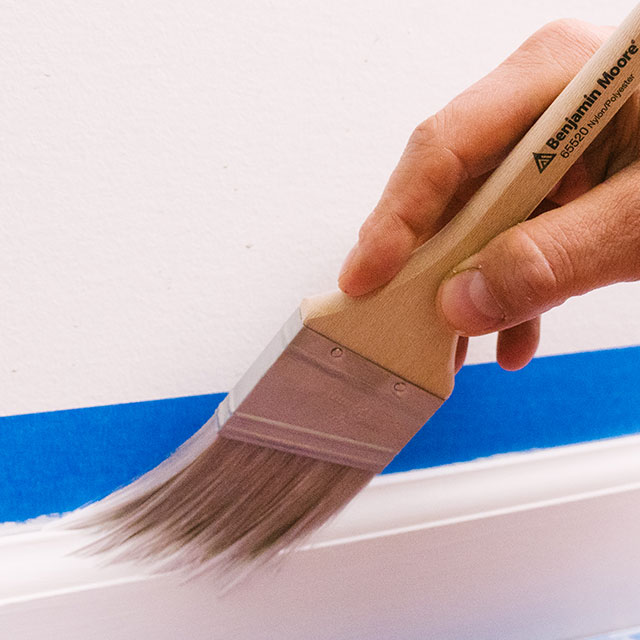 Guided by painter's tape, a homeowner uses a paintbrush to paint trim with white paint. Guided by painter's tape, a homeowner uses a paintbrush to paint trim with white paint.