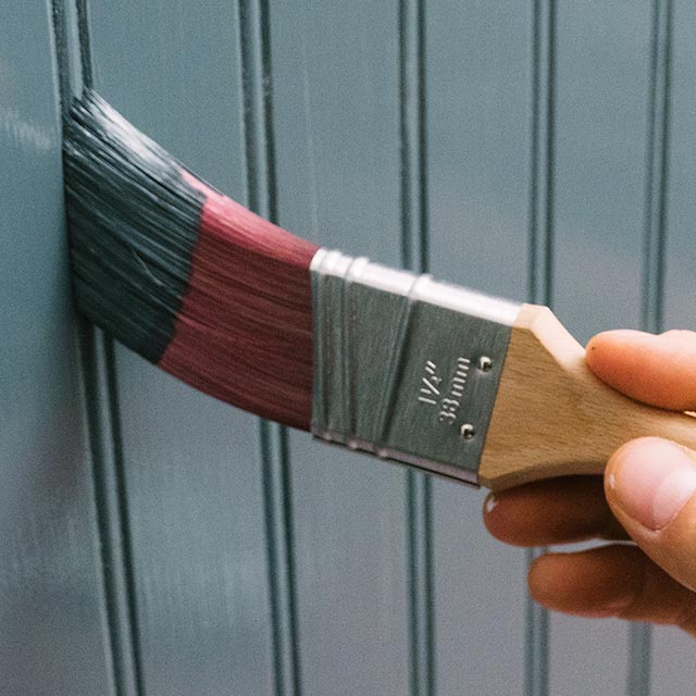 A homeowner using a paintbrush to paint a wall with grooves using a gray-blue paint. A homeowner using a paintbrush to paint a wall with grooves using a gray-blue paint.