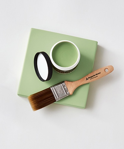 An open Benjamin Moore paint color sample and a paintbrush against a light green canvas. An open Benjamin Moore paint color sample and a paintbrush against a light green canvas.