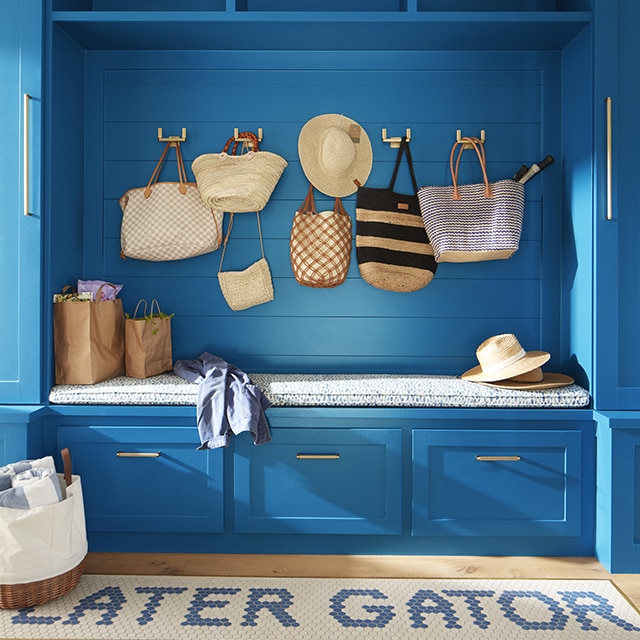 A bright blue-painted mudroom with built-in cabinets, bench and drawers, hanging straw bags and hats, and playful blue and white rug. A bright blue-painted mudroom with built-in cabinets, bench and drawers, hanging straw bags and hats, and playful blue and white rug.