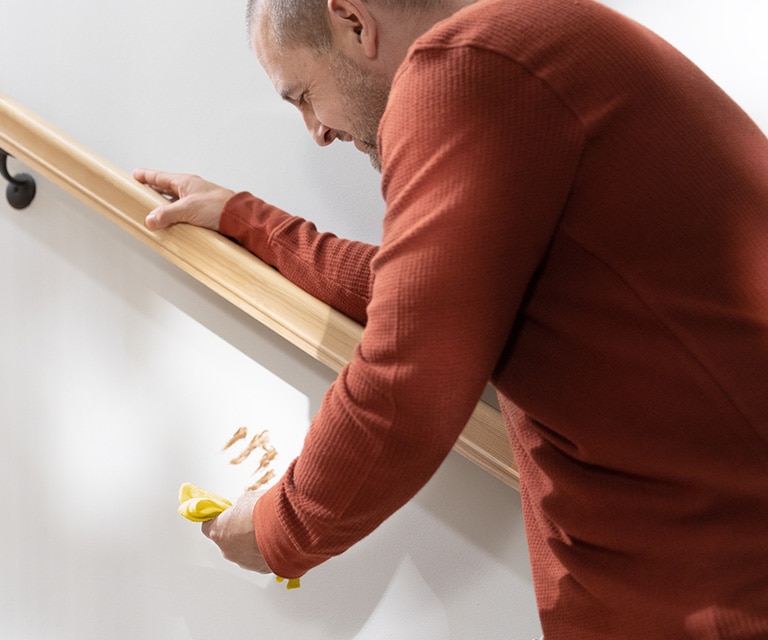 A person in a red shirt leaning against a handrail and holding a yellow cloth to clean a food stain off a white-painted staircase wall. A person in a red shirt leaning against a handrail and holding a yellow cloth to clean a food stain off a white-painted staircase wall.