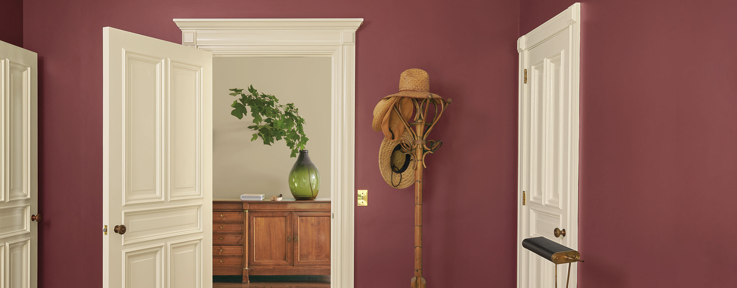 A pretty red painted bedroom with beige trim and doors, a comfy bed with white bedding and an orange pillow, and a coat rack next to an open door looking into a gray painted hallway.