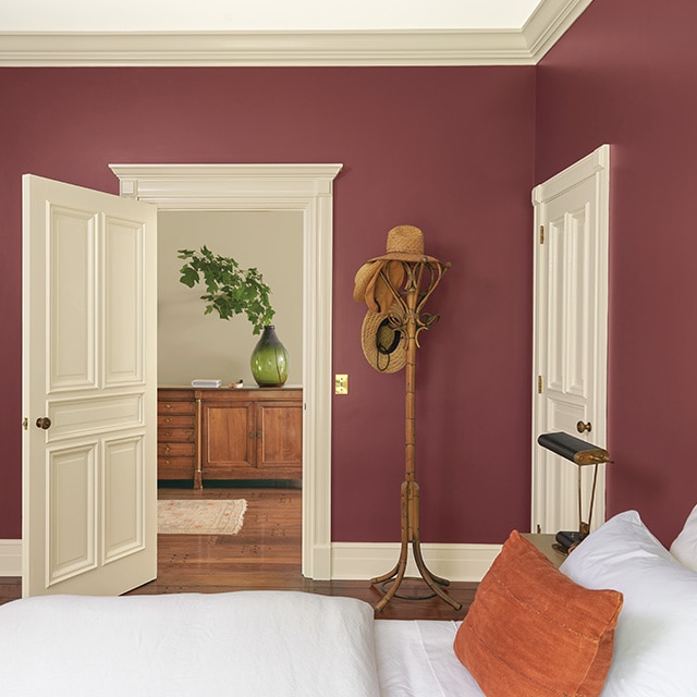 A pretty red painted bedroom with beige trim and doors, a comfy bed with white bedding and an orange pillow, and a coat rack next to an open door looking into a gray painted hallway.