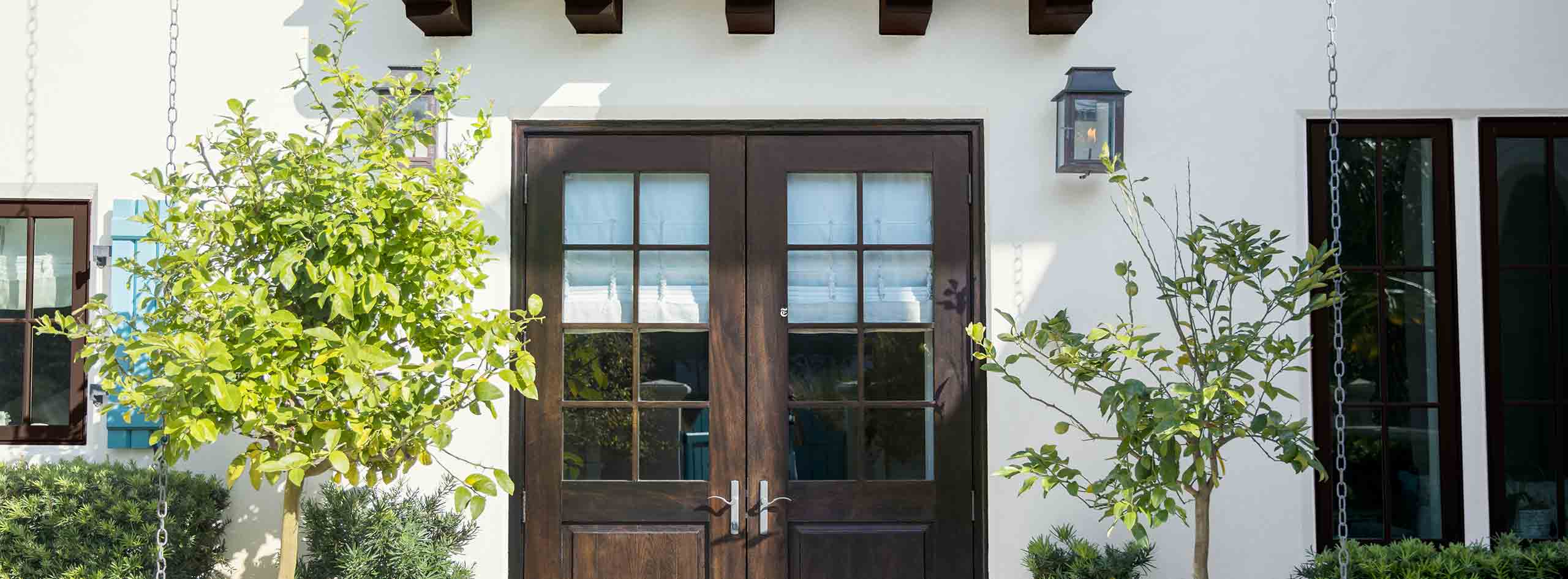 A stucco home with wooden doors and trellis. Bushes and trees adorn the lawn. 