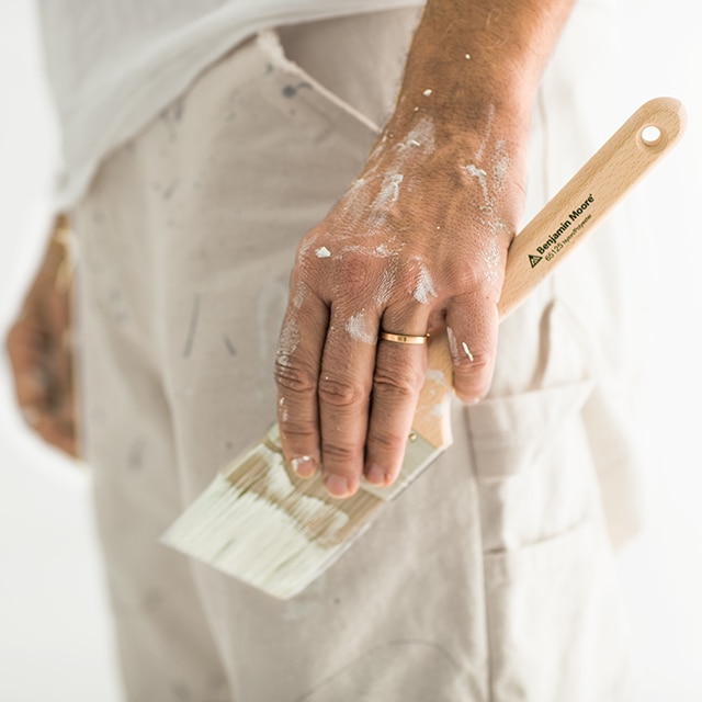 A side shot of the lower half of a painting contractor shows a paint-covered left hand holding a paintbrush. A side shot of the lower half of a painting contractor shows a paint-covered left hand holding a paintbrush.